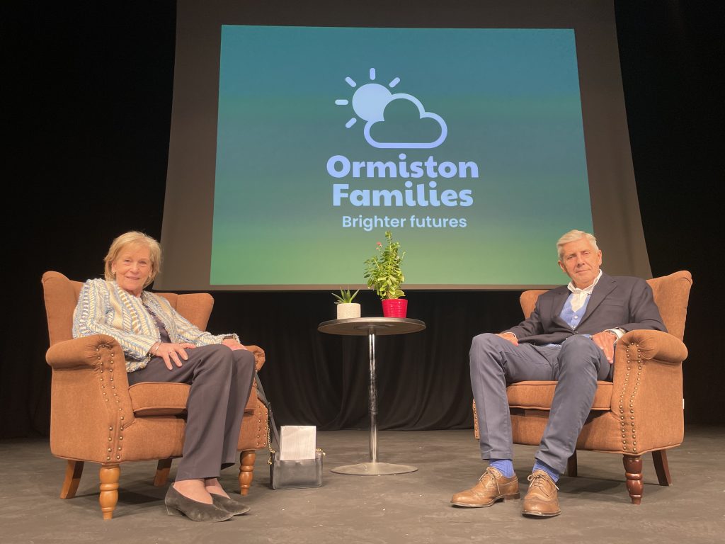 Lady Xa Tollemache and Lord Stuart Rose sit in armchairs on a theatre stage in front of a projection of the Ormiston Families logo.
