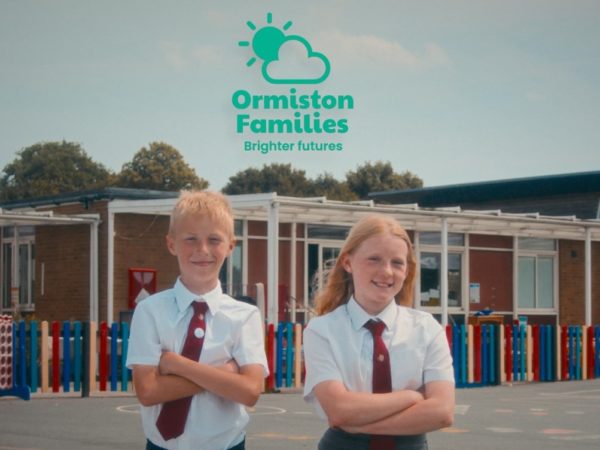Two children pose proudly in front of their school with the Ormiston Families logo above them.