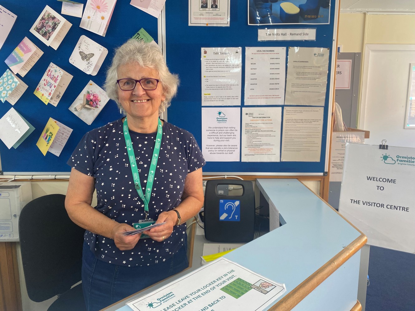 A female charity volunteer with a lanyard round her neck stands in front of the reception counter at a prison visitor centre.