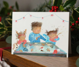 A Christmas card on a table surrounded by festive items - a candle, stockings and holly.