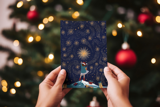 Hands holding a Christmas card with a Christmas tree in the background out of focus.